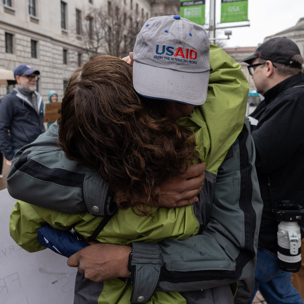 A man in a gray jacket with a U.S.A.I.D. cap hugs a woman in a green jacket.