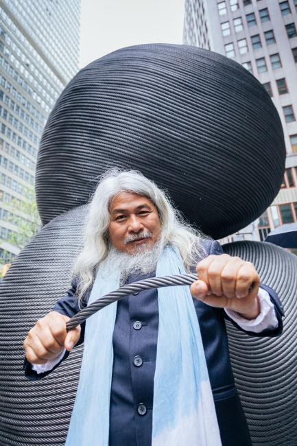 The artist Kang Muxiang in a blue overcoat and light-blue scarf stands beneath his imposing scuplture