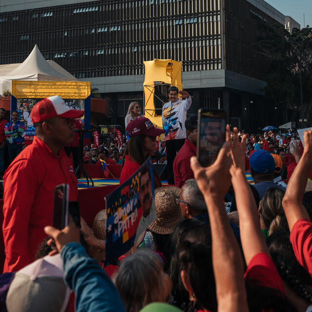 Presidential Nicolás Maduro stands in the background waving to people gathered around him.