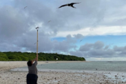 Alphina Luisamoa stands on a beach on a cloudy day holding a rake over her head encouraging a frigate bird to land on it