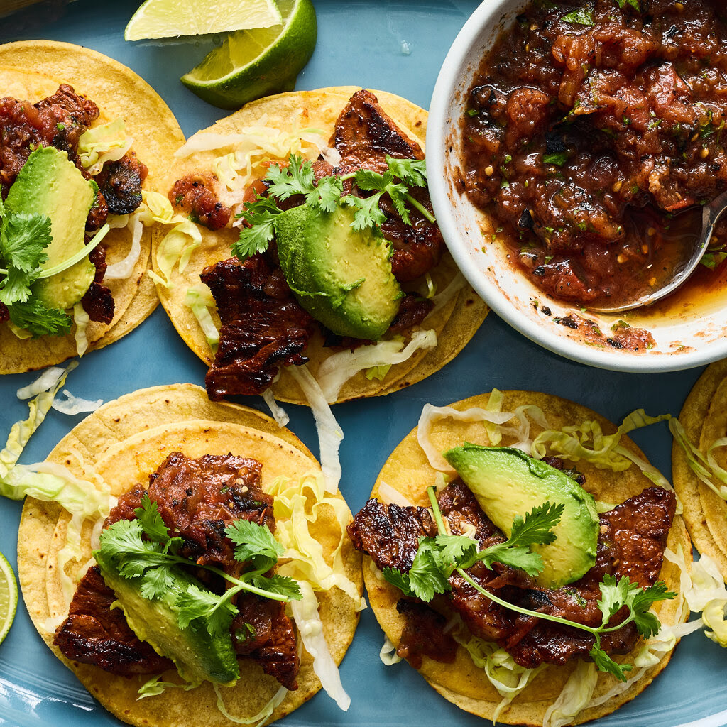 Several tortillas topped with steak, avocado, cilantro and lettuce.