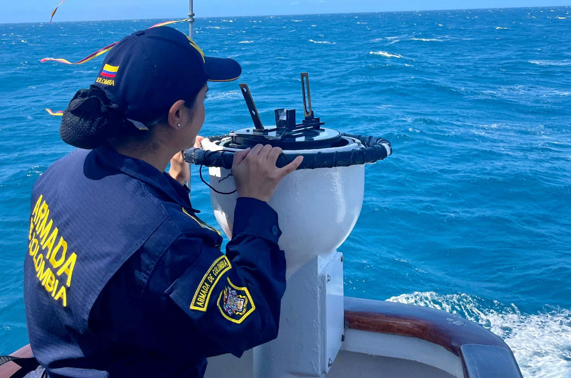 Una mujer vestida con uniforme de la Armada de Colombia está en un barco, mirando a través de un instrumento de navegación. Lleva una gorra de la Armada con una bandera de Colombia en el lateral. El mar azul se extiende en el fondo, con pequeñas olas que sugieren movimiento.