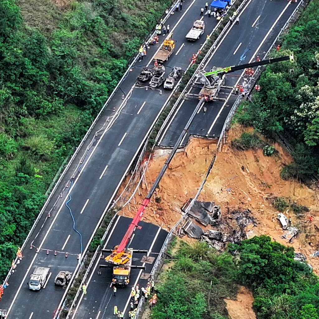 Emergency vehicles, cranes, rescue workers and onlookers are gathered on either side of a large gap in a hillside highway.