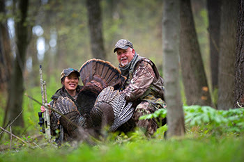 Two hunters pose with a turkey, smiling.