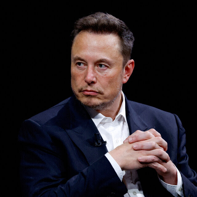 Elon Musk, wearing a suit with no tie, sits onstage with his fingers interlaced.