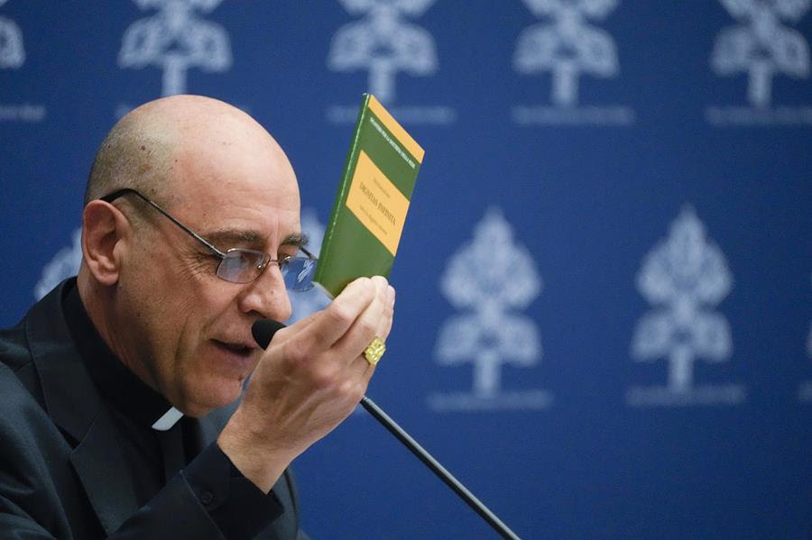 Cardinal Victor Manuel Fernandez speaks into a microphone while holding up the "Infinite Dignity" document.