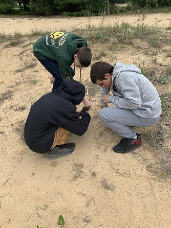 On a sandy shoreline, three young men kneel in a circle to examine a small patch of vegetation.