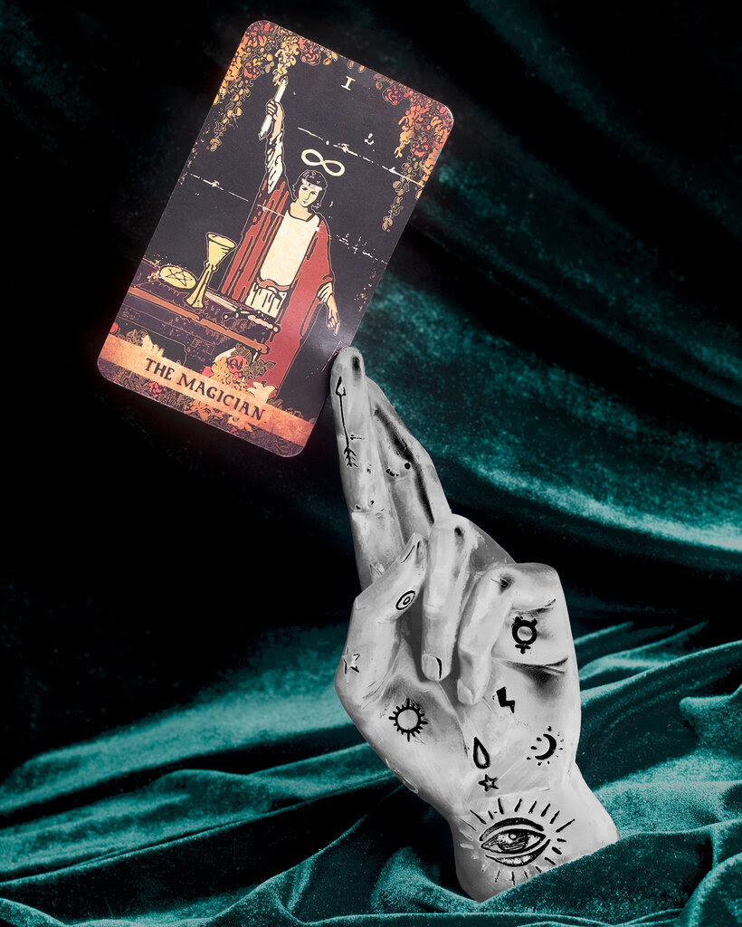 A photo illustration of a tattooed hand holding a tarot card that says: “The Magician.”