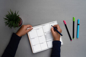 Journaling daily for mental well being and personal development. Weekly planner set up on a minimal grey table top, with sketch pens and a green house plant