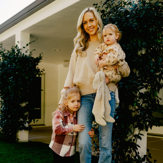 A blonde-haired woman stands outside a building, in front of a bush, smiling with two young children. One stands by her leg, looking, at the camera. The other is in her arms. 