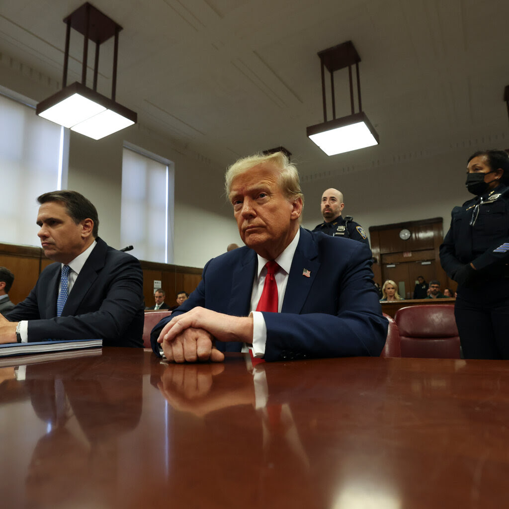Donald Trump, in a blue suit and red tie, sitting in a courtroom.