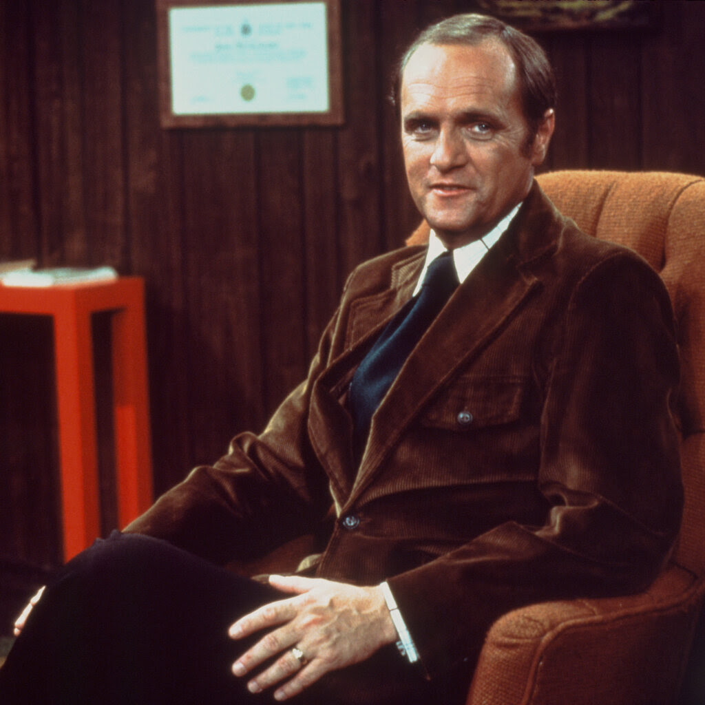 Mr. Newhart in a brown blazer, white shirt and blue necktie while sitting in an orange upholstered chair in a paneled room and looking at the camera with a slight smile.