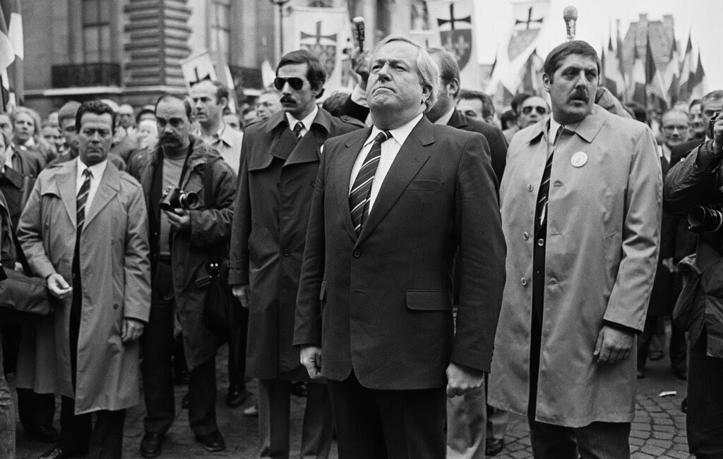 A black and white photo of a gray-haired man standing on a city street — his shoulders thrown back, his chin up — with a crowd behind him, including three men close by in trench coats. He wears a dark suit, white shirt and striped necktie. 