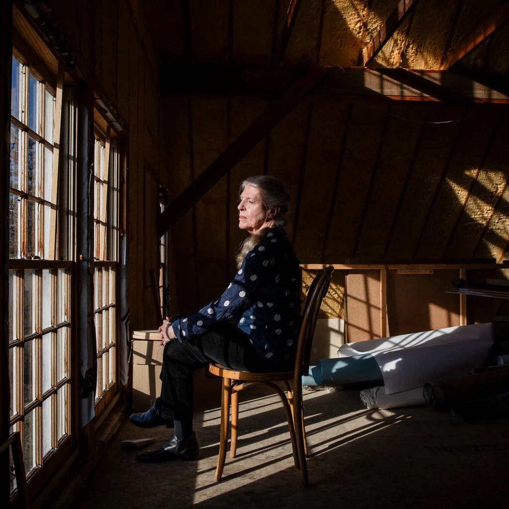 An image of a woman sat on a chair in a darkened room, facing the window. 