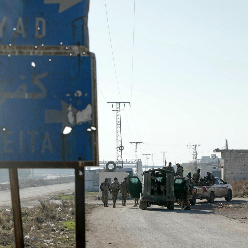 Soldiers stand around trucks on a dusty road with power lines in the distance and a rusted road sign with bullet holes in the foreground.