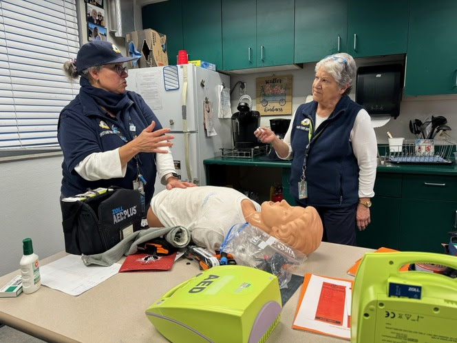 Two ferry employees speak to each other with a CPR manikin and an automated external defibrillator on the table in front of them