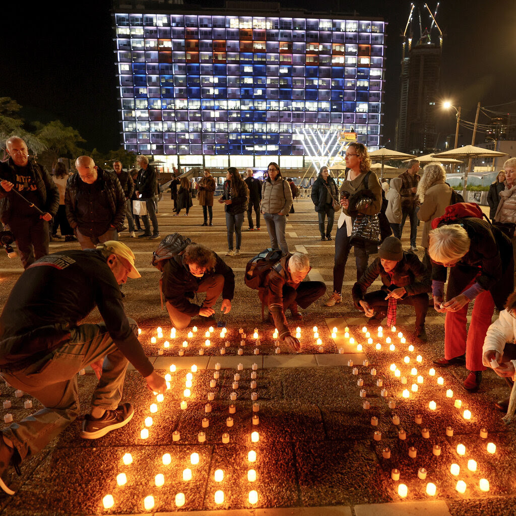 People leaning over a grid of candles on the ground in a city square near a brightly lit building at nighttime. 