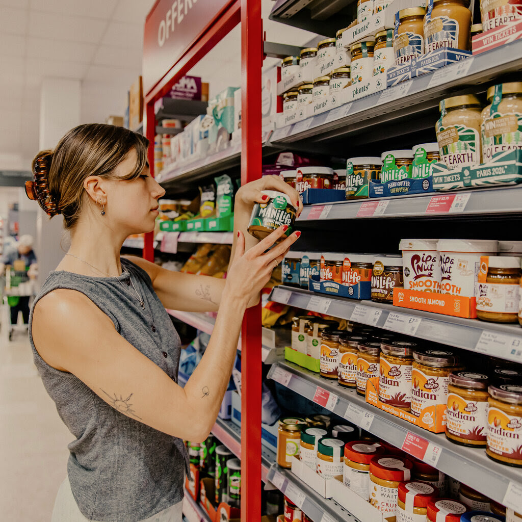 A woman holds a jar of ManiLife peanut butter in a British grocery store in front of an array of other jars of peanut butter.