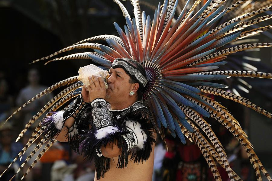 A member of the Grupo Coatlicue, wearing traditional clothing including a large feathered headdress, blows on a conch shell.