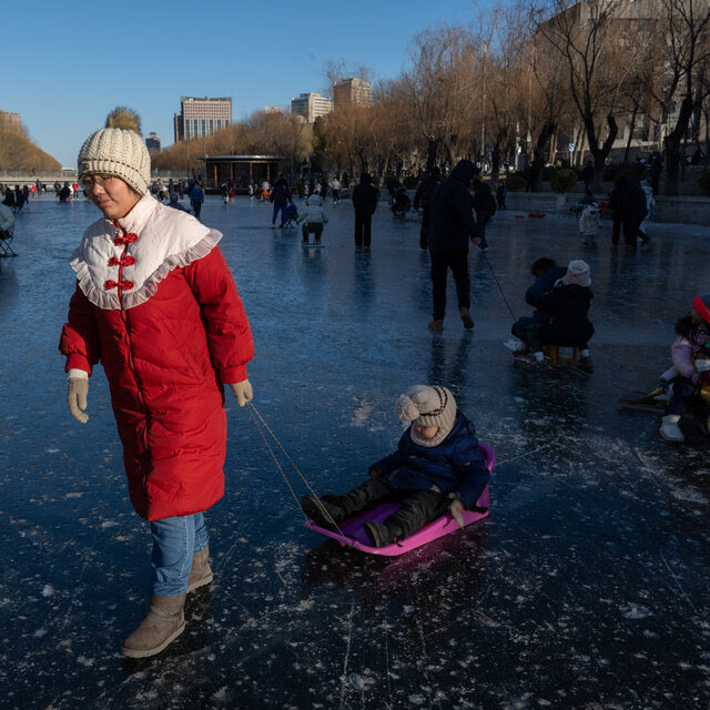 On a frozen river crowded with people, an adult wearing a heavy red coat pulls a child in a pink sled across the ice.