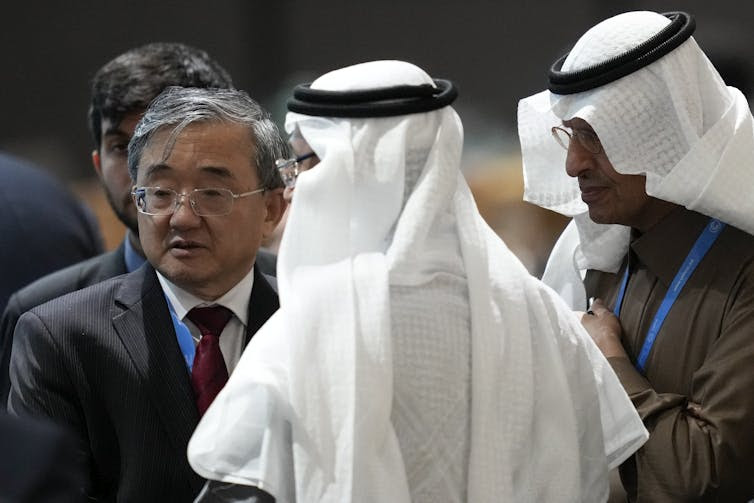 An Asian man stands next to two Saudi officials in white head scarves.