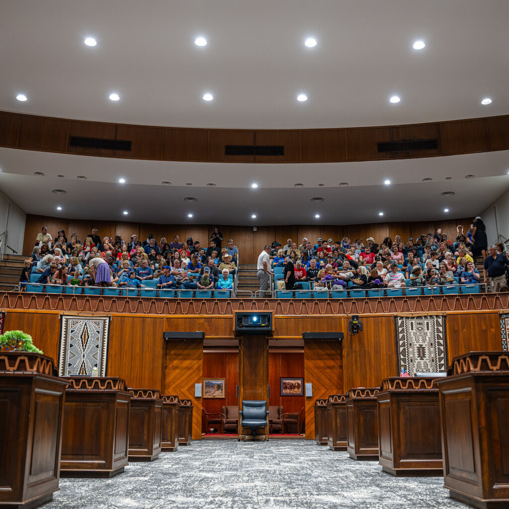 A view from between rows of desks in a legislative chamber. Above, people sit in a raised visitors’ gallery.