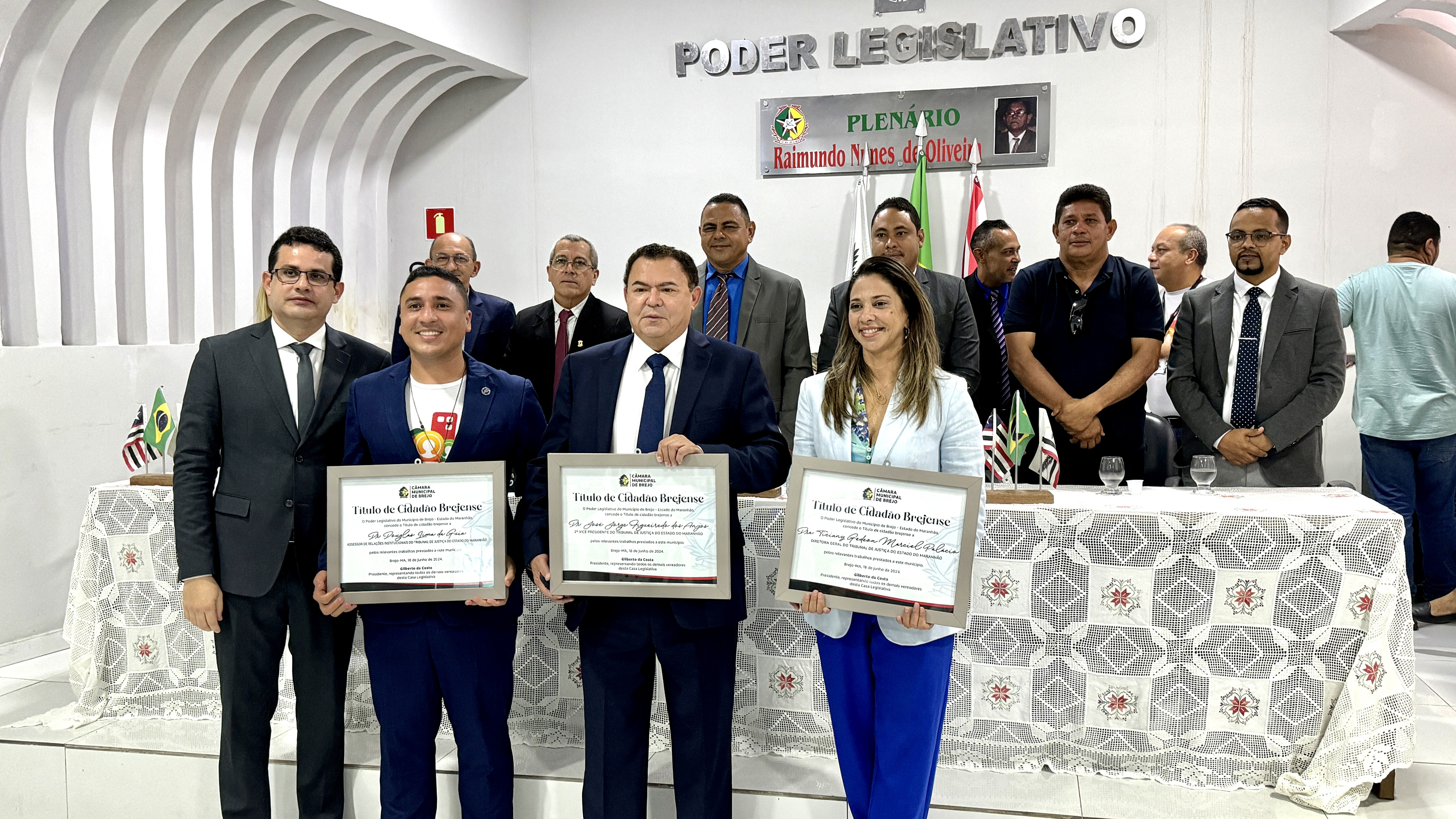 A imagem mostra um grupo de pessoas em um ambiente formal, possivelmente uma câmara legislativa ou governamental, conforme indicado pela placa ¿PODER LEGISLATIVO¿ ao fundo. Os indivíduos estão vestidos com trajes de negócios e três deles seguram certificados, sugerindo uma cerimônia de premiação ou evento de reconhecimento formal.