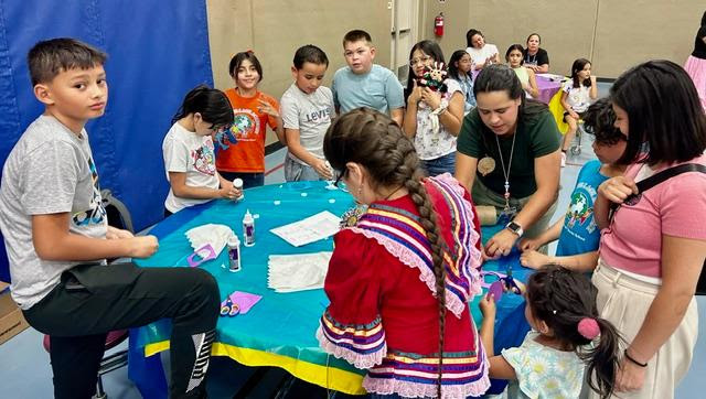 A group of children and adults gathered around a table engaged in arts and crafts one child wearing traditional clothing colorful supplies and decorations on the table smiles and concentration as they work