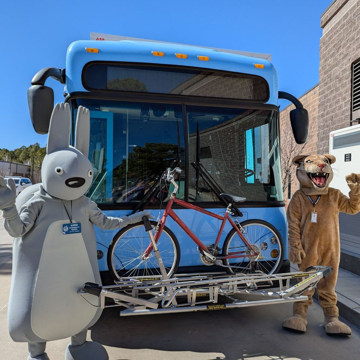 Sustainability office mascot Albert and Mountain Line Mascot pose in front of bike mounted on the bus