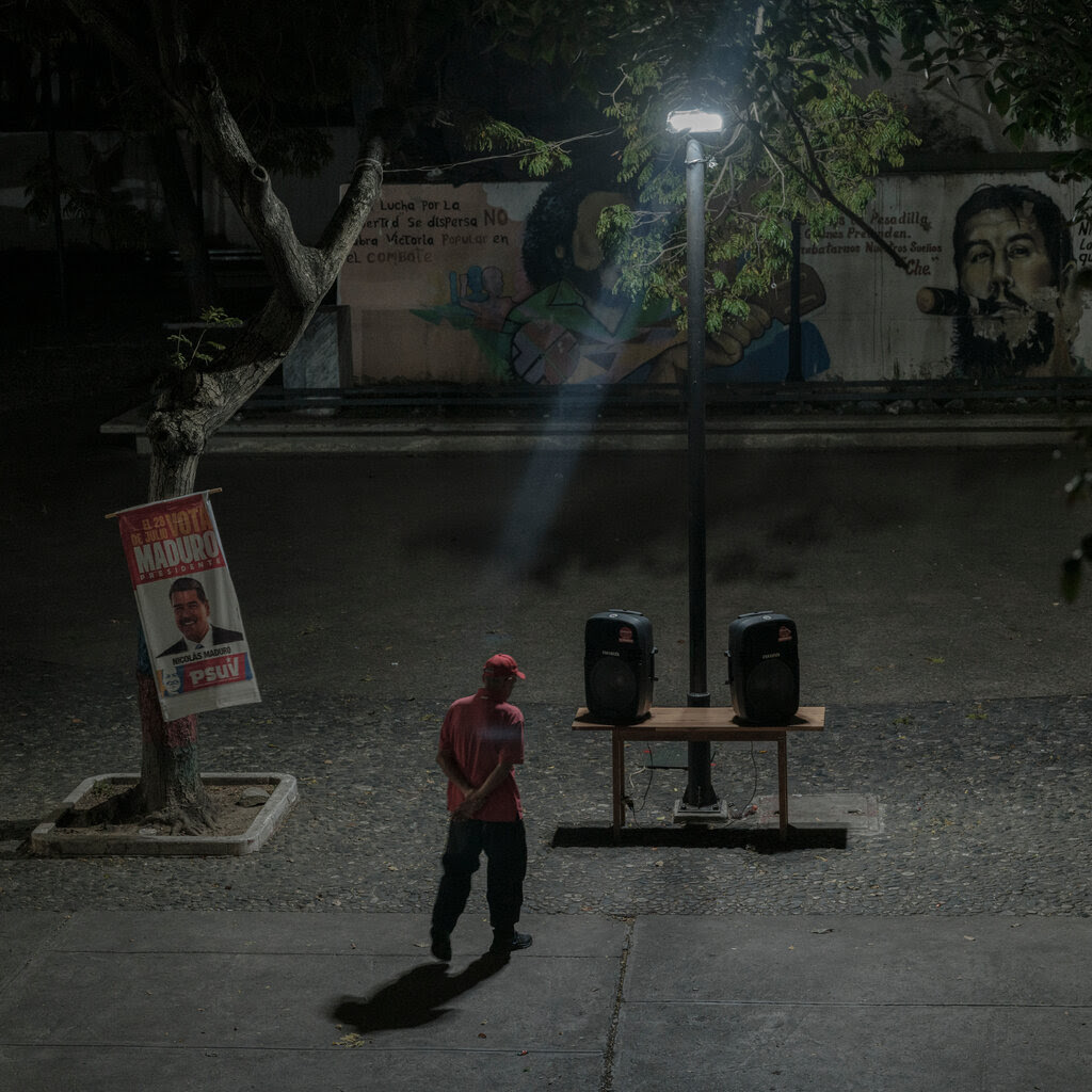 A man stands in front of speakers placed on a table outdoors at night. A campaign poster for President Nicolás Maduro of Venezuela is attached to a tree.