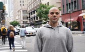 Man in sweatshirt smiling, standing on the sidewalk of a bustling city.