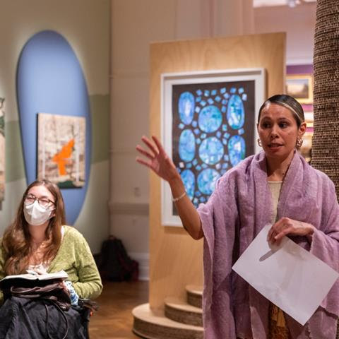 A woman stands speaking in an exhibition space.