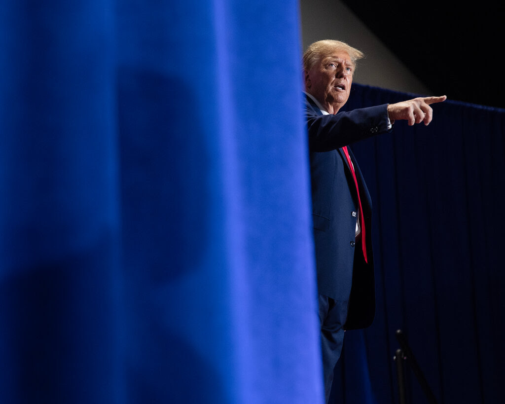 Donald Trump pointing with his right hand and wearing a dark suit. He is standing near a blue curtain.