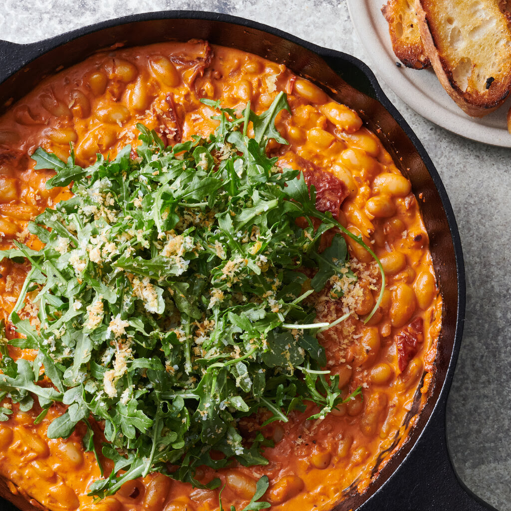 A cast-iron skillet holds creamy spicy tomato beans and greens. Toasted slices of bread are on a small dish nearby.