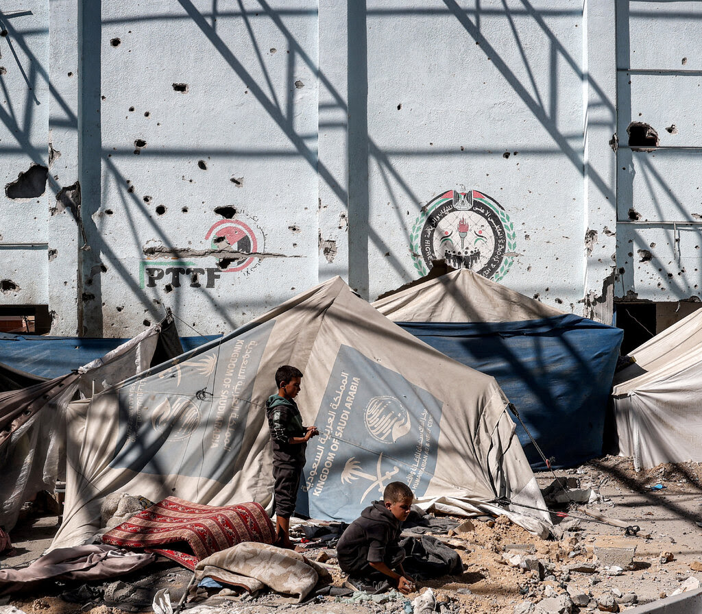 Tents stand close together alongside a pockmarked wall, with two children in the foreground amid debris.