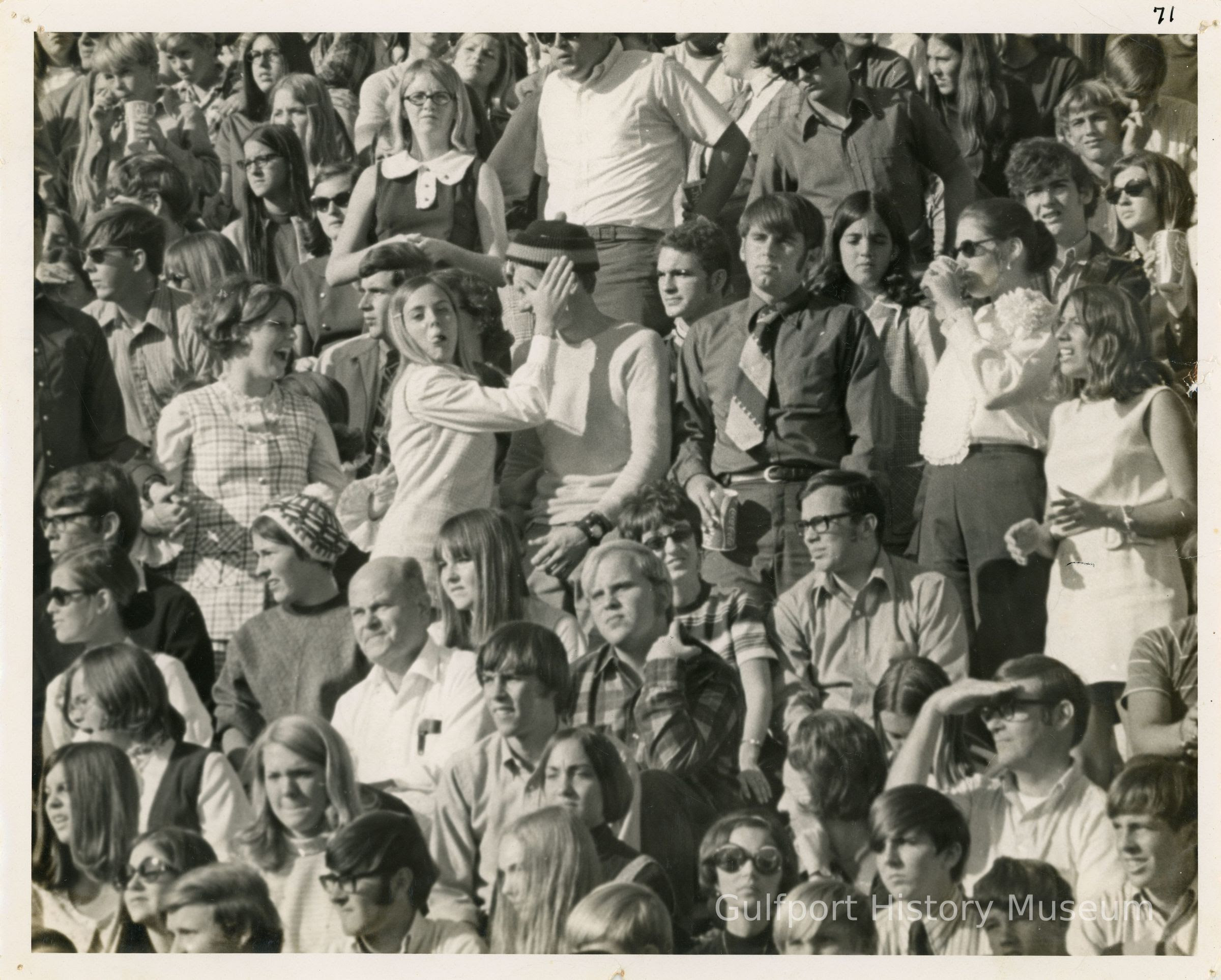 A black and white photograph of a crowd of high school students. Most of them are facing the left side of the photograph, while some are looking at the camera. In the center of the crowd, a girl is covering someone's face with her hand. Another girl is standing next to them and laughing. 