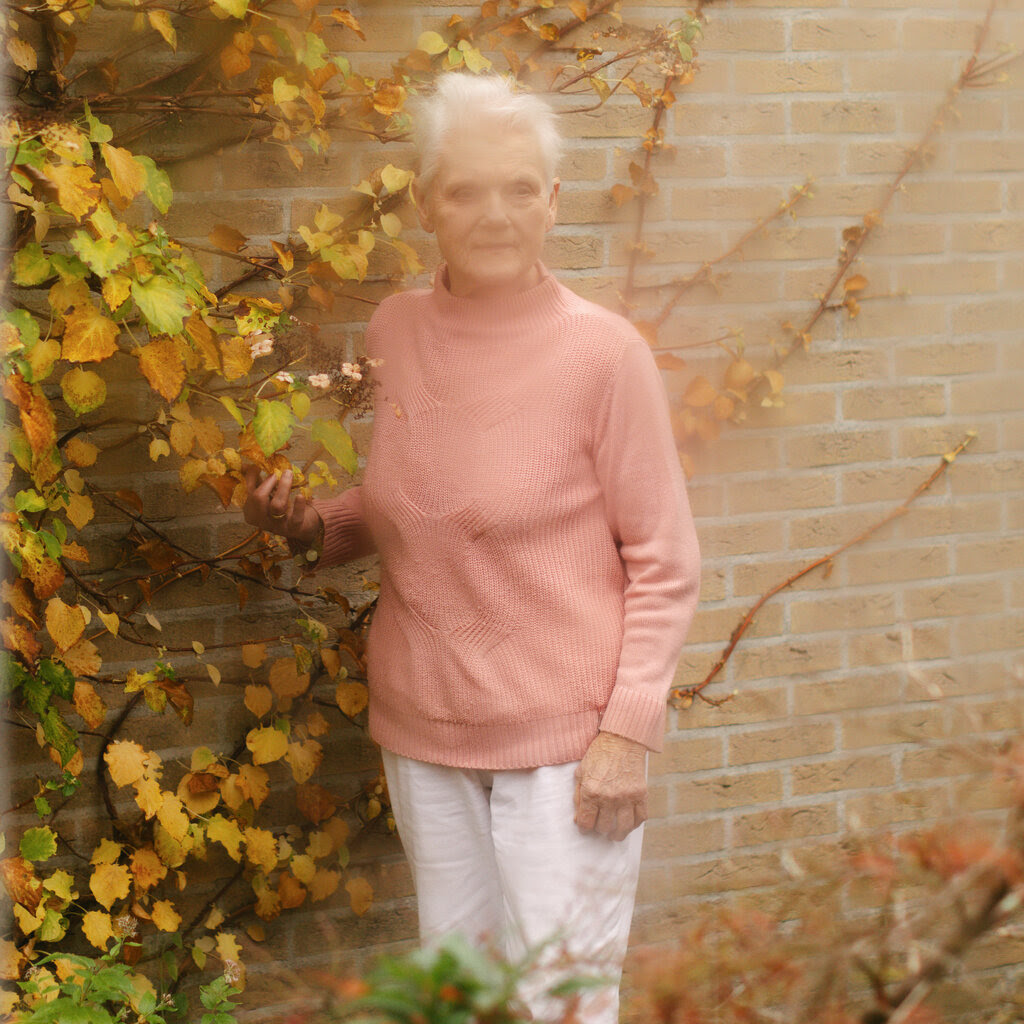 A woman in a coral-colored sweater standing in front of a leaf-covered brick wall.