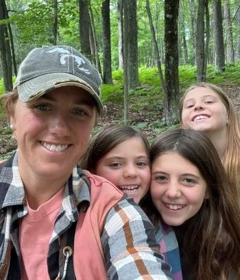 Smiling woman in slate blue baseball cap with Michigan state outline, surrounded by three smiling young girls. Thick forest in background