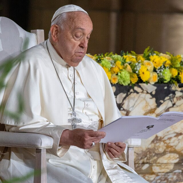 Pope Francis sitting and reading papers with flowers in the background.