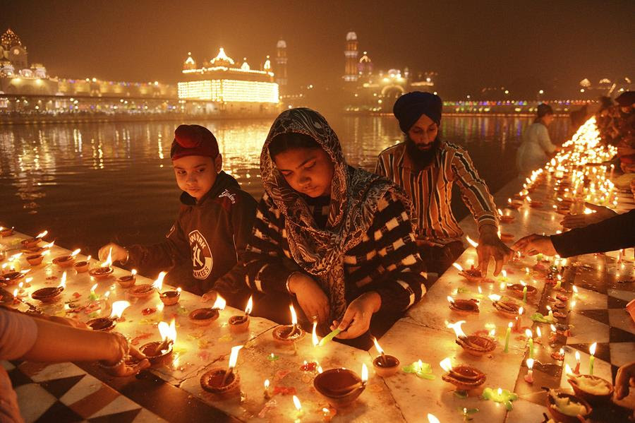 Sikh devotees light clay lamps next to a pond. The walls lining the pond are filled with many small clay lamps that give the scene a warm tone.