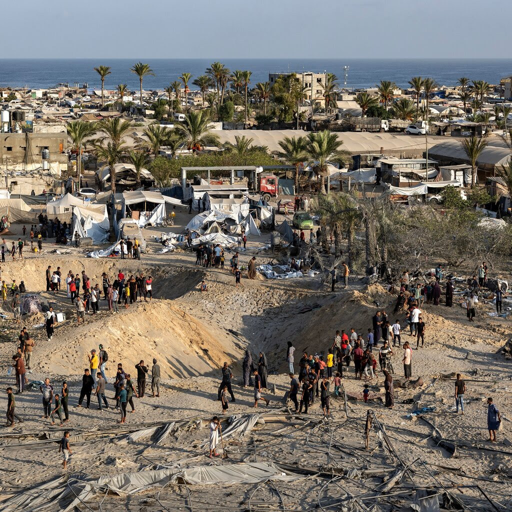 People cluster around two large craters in a sandy area surrounded by tents and low-rise buildings near the seaside. 