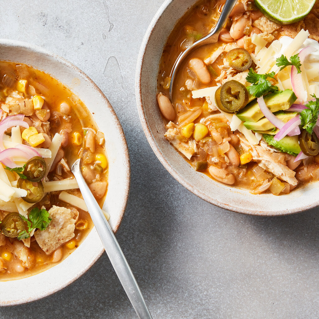 Two servings of white chicken chili are served in white bowls and topped with shredded cheese, slivered red onion, avocado slices, crushed tortilla chips and limes for squeezing.