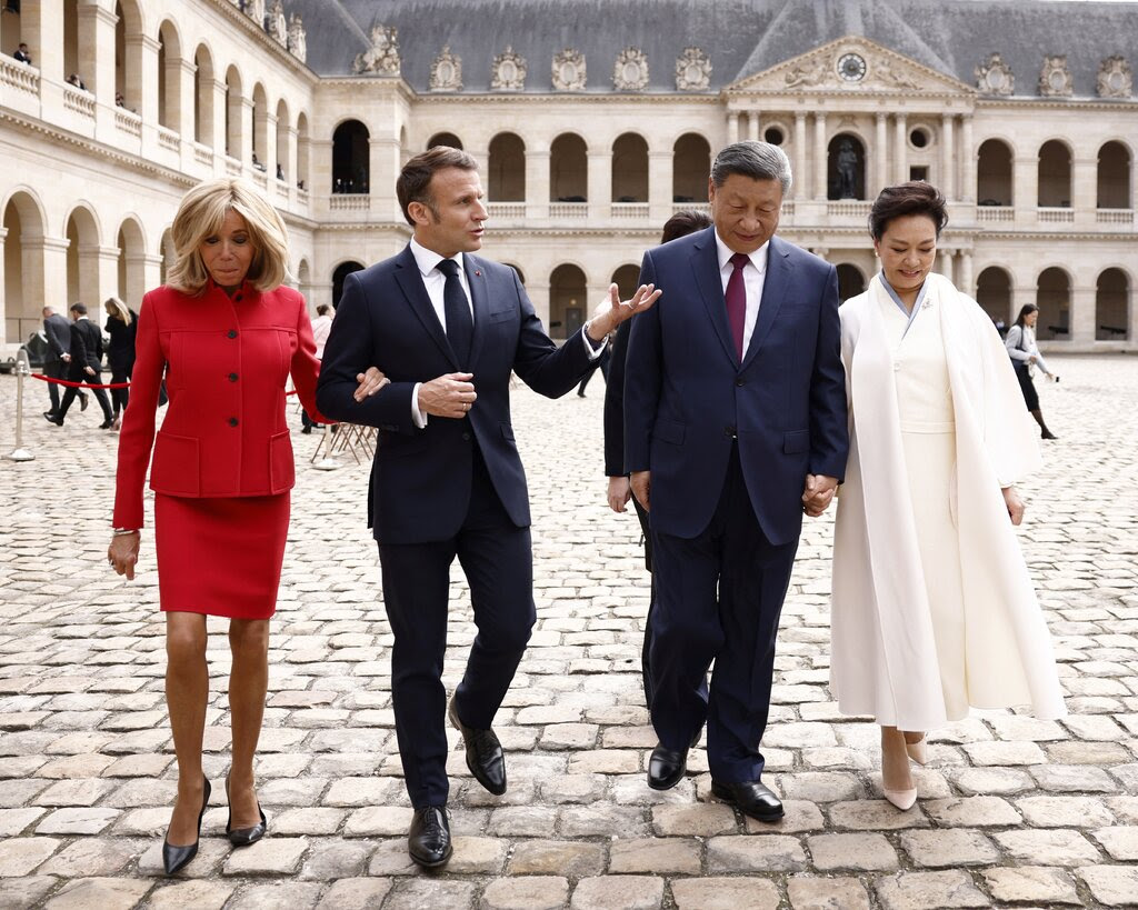 Four people walking -- a woman in a red dress is holding the arm of a man in a suit. He is gesturing to another man in a suit, walking with a woman in white. 