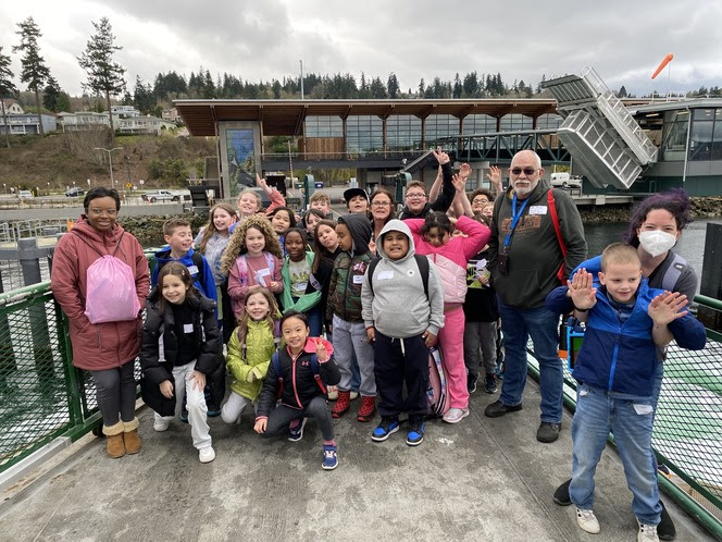 Several children and a few teachers posing for a photon on the outdoor deck of a ferry with Mukilteo terminal in the background