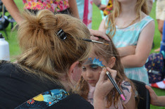 a young girl is having face paint applied to her by a female adult, back facing the camera