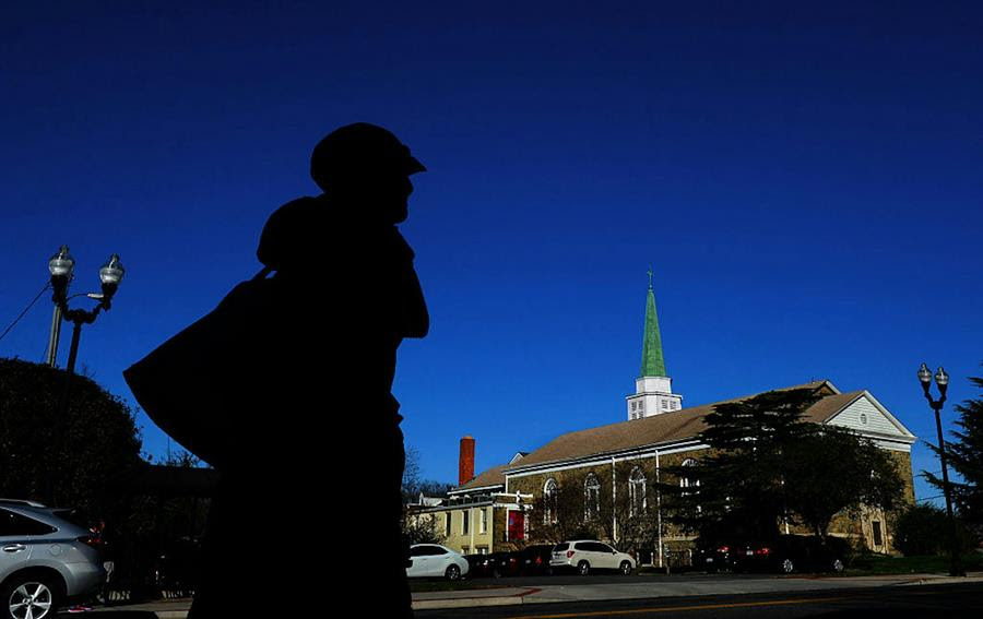 A person in a hat and coat, silhouetted against a dark blue sky, walks across the street from a church.