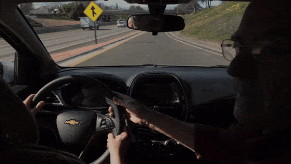 A GIF shows a man in the passenger seat of a car holding the wheel with one hand to steady it, as the driver grips the wheel with two hands while learning to drive.
