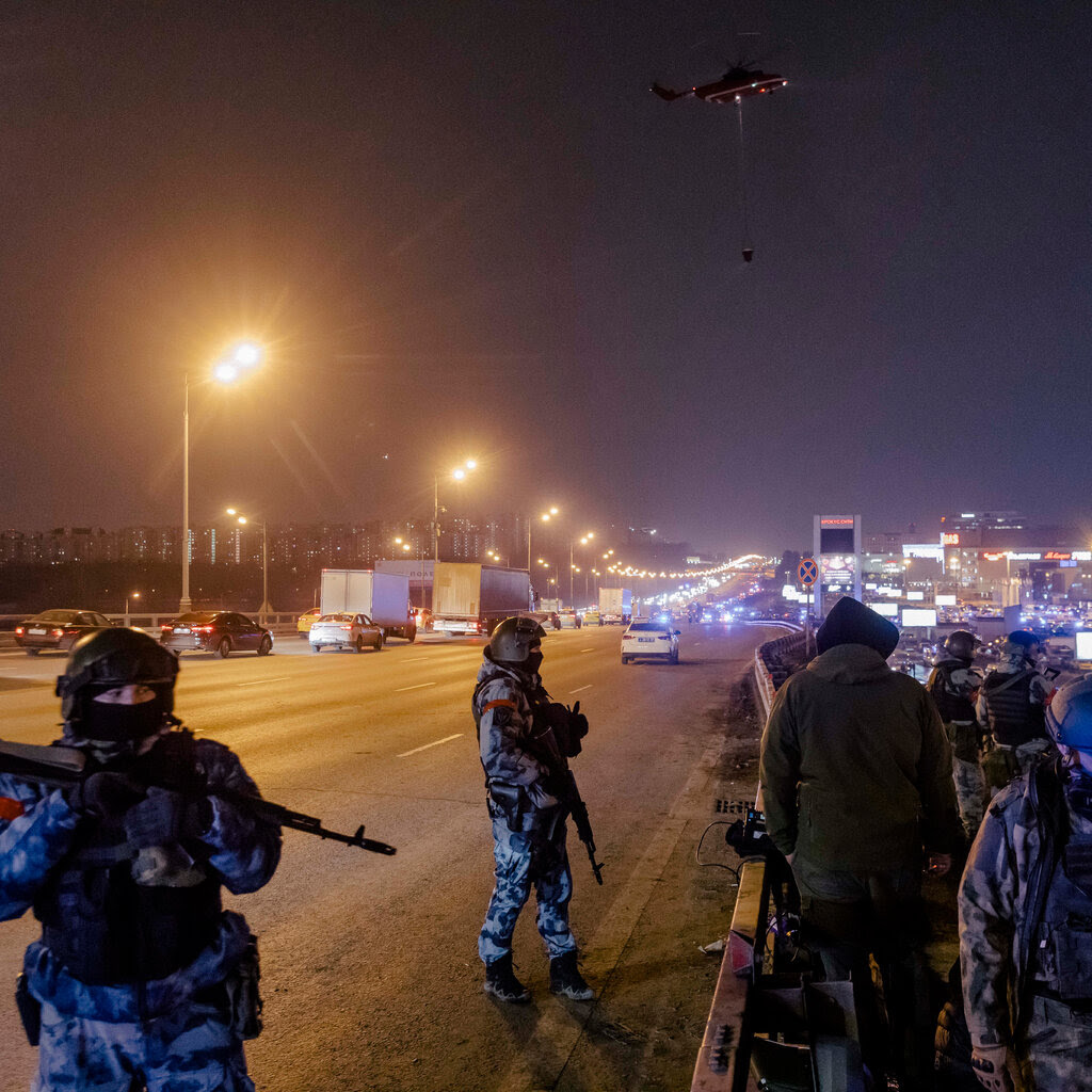 Masked, heavily armed security personnel standing on a large road at night.