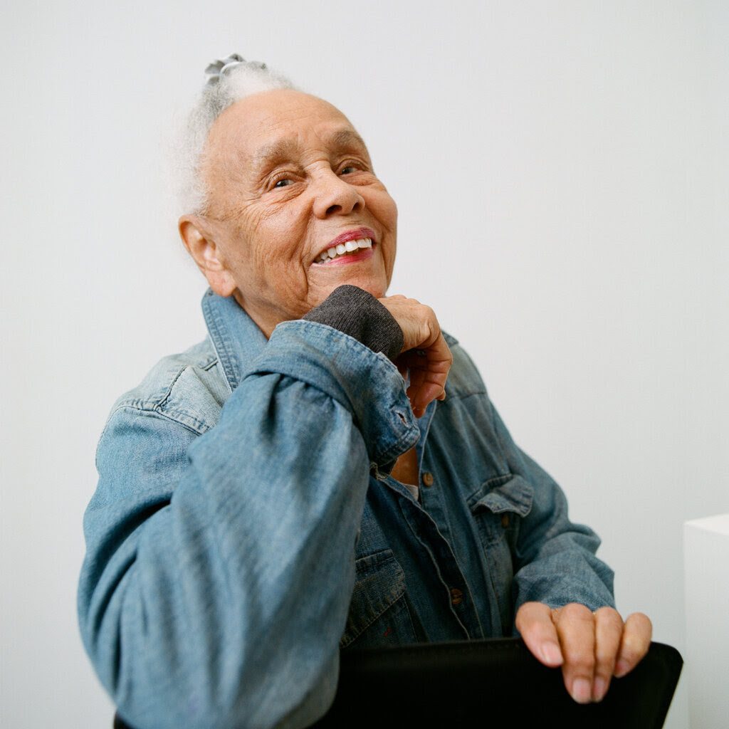 Betye Saar, wearing a denim jacket, looks over her shoulder and smiles. She is wearing a light red lipstick and her gray hair is tied up on top of her head.