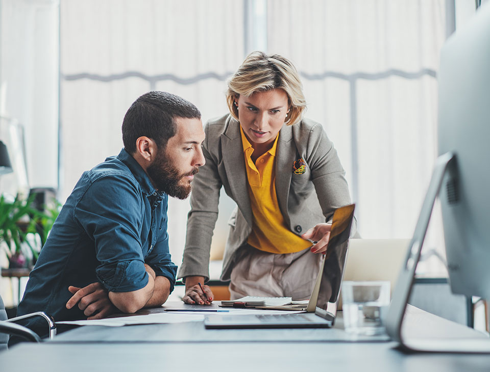 The image depicts a professional woman and man intensely reviewing documents and a computer screen in a modern office setting, suggesting a collaborative discussion or review process.